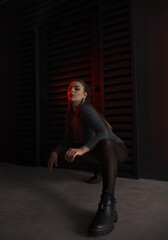 teen fashion. young brunette girl in a shiny silver bodysuit and boots sits fashion on floor with hands on the knees near black wall background in studio with red light. fashion concept, free space