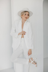 Confident young blond woman smiling, looking at camera isolated on white background. Studio portrait of successful friendly female in white suit and hat, posing over white wall.