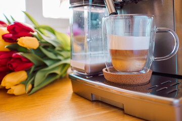 Cup of coffee with milk and tulips flowers on wooden kitchen table. Freshly brewed cappuccino espresso in coffee machine for breakfast. Stylish home interior decor. High quality photo