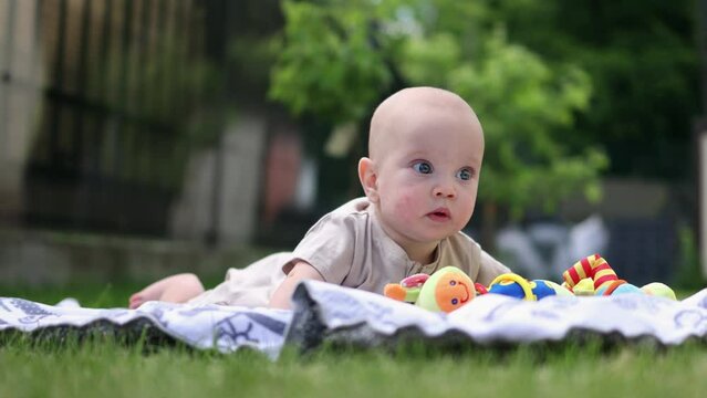 cute little baby infant lying on tummy on grass outdoors in garden playing with colorful toy. tiny 4 months child kid looking around, exploring green nature. caucasian baby on stomach, warm sunny day