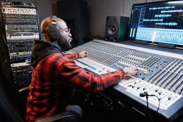 Stylish African American music producer wearing headphones sitting at mixing desk recording song in...