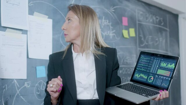 Caucasian blonde businesswoman standing at blackboard and holding laptop while comparing data from stuck paper sheets on board with graphs on screen. Business, work concept