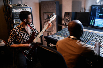 Stylish young Black musician playing electric guitar while music producer working on mixing console...