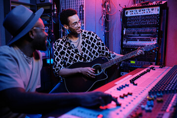 Mature Black music producer and young guitar player enjoying working on album in recording studio,...