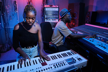 Modern young African American female musician playing digital keyboard while sound engineer working...