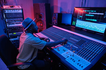 Stylish mature African American man wearing hat creating soundtrack using mixing console in...