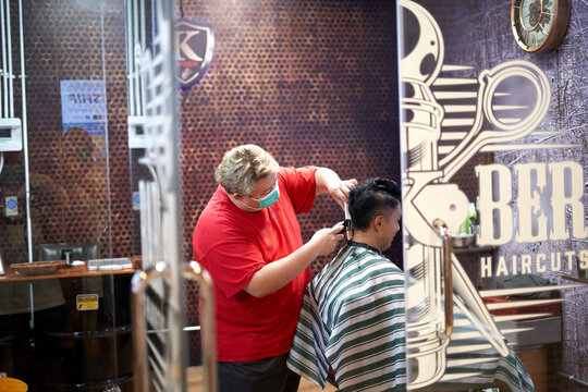 Fat barber standing cutting the hair of an asian client in a barber shop.