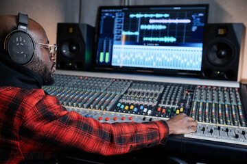 Fototapeta na wymiar Modern African American sound engineer wearing headphones sitting at mixing console working on music track