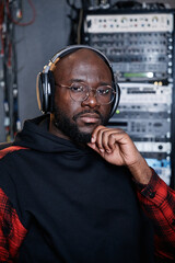 Portrait of modern African American man with beard on face wearing headphones and eyeglasses sitting in recording studio looking at camera