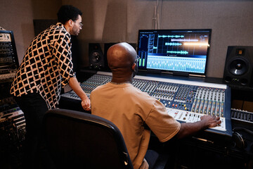 Mature African American producer and young singer creating music track using mixing console in...