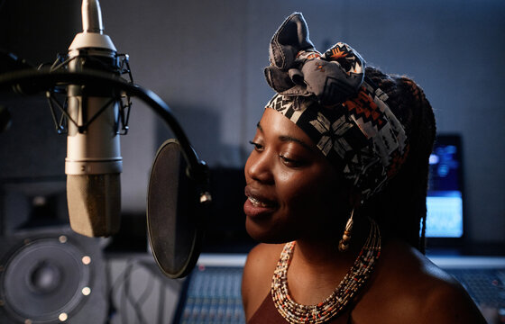Portrait of stylish young African American female singer standing in front of microphone singing song in recording studio