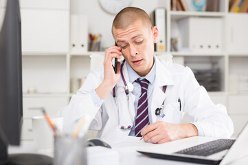 Male doctor working in the clinic consults a patient on the phone, sitting at a desk in the office