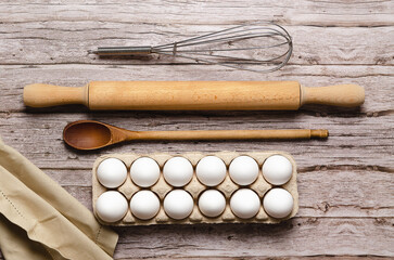A whisk, a rolling pin, a wooden spoon, some white eggs in an egg container and a piece of cloth on...