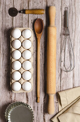 A whisk, a rolling pin, a wooden spoon, some white eggs in an egg container and a piece of cloth on a wooden table. 