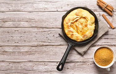 Apple galette in a black pan, brown sugar in a cup and cinnamon sticks, on a wooden table. 