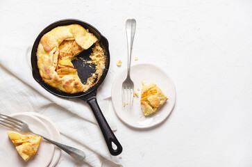 Apple galette in a black pan, two pieces of galette in white plates with forks and a piece of white cloth on a white background. 