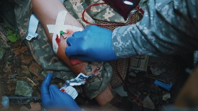 Military medic receiving blood transfusion provides first aid to a soldier who inserts a needle into a vein. Hands