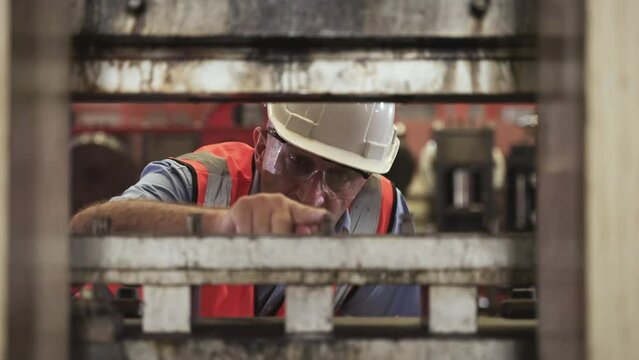 Professional heavy industry senior engineer man in uniform wearing safety goggles and hardhat with accuracy checking of mold die on hydraulic press machine in factory. Male maintenance worker concept.