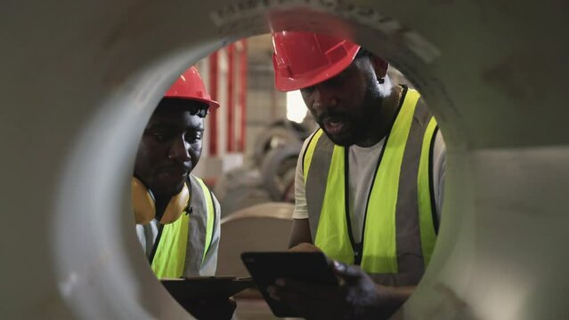 Two African man workers in uniform wearing safety hard hats holding tablet and clipboard inventory checking stock in warehouse at factory industry. Professional engineer inspection roll of metal sheet