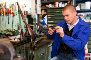Fototapeta na wymiar Portrait of focused skilled craftsman of weapons workshop engaged in shotgun repairing, using vernier caliper to measure parts size