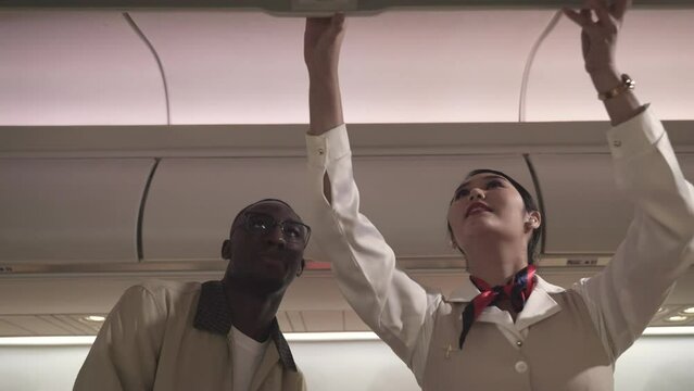 Asian female flight attendant or airplane stewardess in uniform with help service for African man customer keep suitcase to overhead luggage compartment on cabin airplane. Business airline concept. 