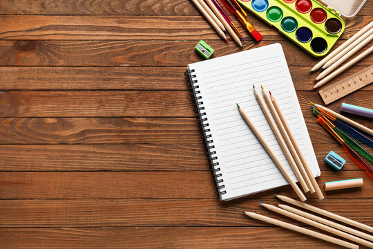 Colorful pencils, blank notebook and paints with brushes on wooden background