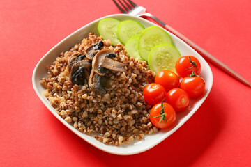 Plate of tasty buckwheat porridge with mushrooms and vegetables on red background