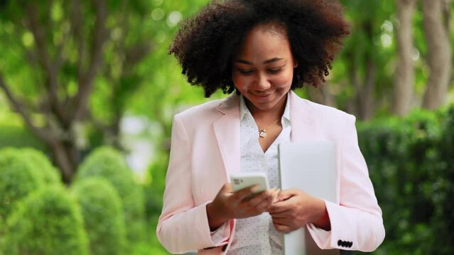 Smiling  happy  young  African American business woman with Afro hair holding smart phone, checking social media, using mobile app  walking at outdoor.  