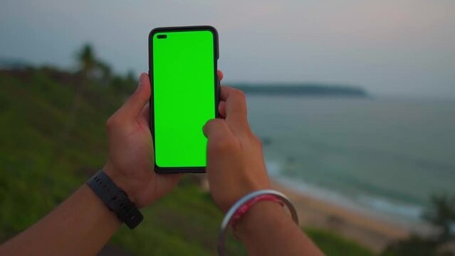POV shot of hand holding mobile with green screen in front of the sea during the sunset. Chroma key mock-up on smartphone in hand at the sea shore in Goa, India. Green screen mockup template of Mobile