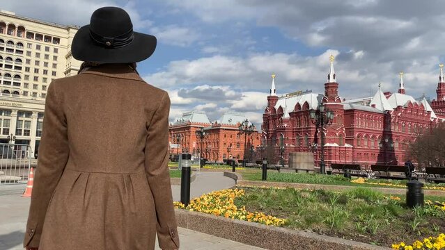 Young beautiful lady elegance madame in hat and coat walking on Red Square  near Historical Museum, Kremlin, Alexander Garden and Okhotny Ryad. Spring, blooming flowers. Center of Moscow, Russia.