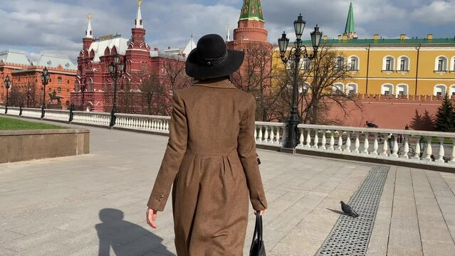 Young beautiful lady elegance madame in hat and coat walking on Red Square  near Historical Museum, Kremlin, Alexander Garden and Okhotny Ryad. Spring, blooming flowers. Center of Moscow, Russia.