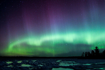 Northern lights erupt over a lake in Minnesota in the dark sky overhead shining a rainbow of light and colors over the frozen water and forests