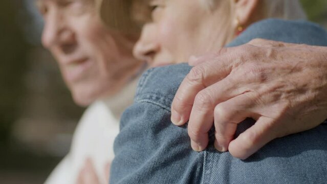 Closeup shot of senior couple enjoying warm autumn day in park. Loving elderly husband embracing his wife while couple relaxing and talking outdoors. Love, retirement concept