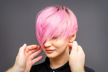 Hairdresser with hands is checking out and fixing the short pink hairstyle of the young white woman in a hair salon