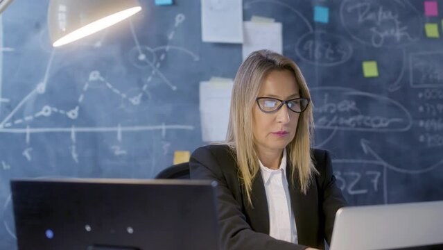 Beautiful Caucasian businesswoman working on laptop and making notes, then taking data sheet from board and comparing information. Blackboard with charts and stickers in background. Business concept