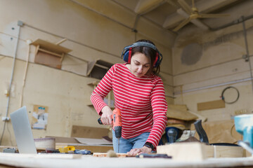 A female carpenter in a striped red shirt stands in her workshop and drills a hole with an electrical drill