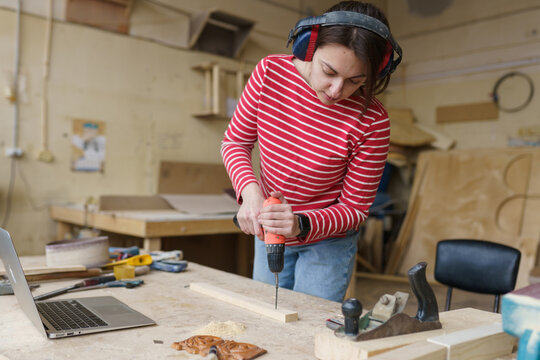 A female carpenter in a striped red shirt stands in her workshop and drills a hole in a wooden plank electric drill