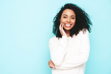 Beautiful black woman with afro curls hairstyle. Smiling model dressed in white summer dress. Sexy carefree female posing near blue wall in studio. Tanned and cheerful