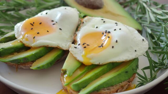 Modern Two Toasts with avocado and fried egg Spreading Yolk, rosemary 4K. Preparing healthy toast breakfast avocado slices rye wholegrain bread, sunny side up egg on top. breakfast for two, couple
