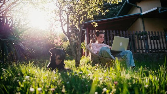 A young adult woman working on her laptop while sitting in the garden of her village house and her dog is lying on the lawn next to her.