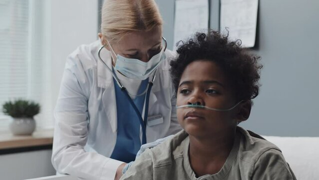 Medium of ill Black boy with oxygen tube sitting on bed in clinic room at daytime, Caucasian female pediatrician in medical mask and gown using stethoscope on his back, giving him instructions