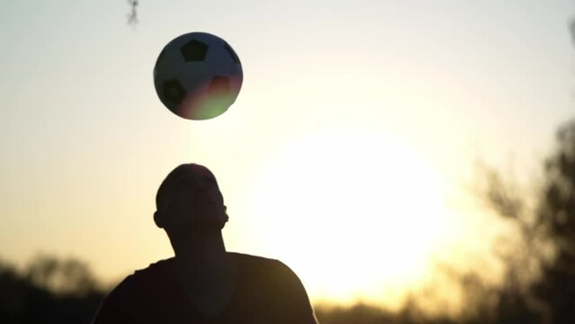 Man Kicks Soccer Ball With His Head. Silhouette of Young Man Kicks Football Ball with His Head on Background of Sky and Setting Sun. Slow Motion