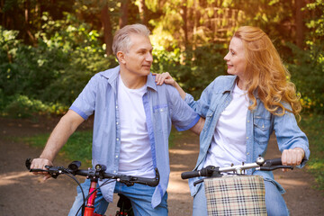 Cheerful active senior couple with bicycle walking through park together. Perfect activities for seniors in retirement lifestyle wearing casual wear smiling happily