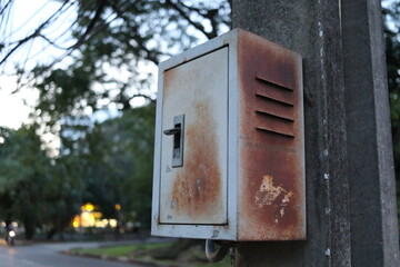The old metallic box is hanging on cement pole in public park. Outside of box has brown rust.