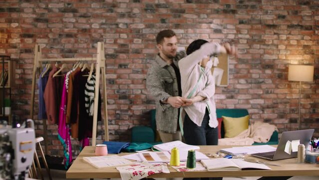 In a designers space two people, a woman with pixie hair and a man with a beard are dancing around with eachother and having fun as they re taking a break from their work