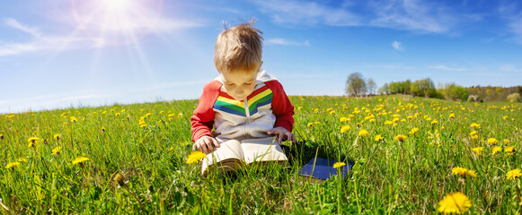 Little child sitting with book on the field with dandelions.
