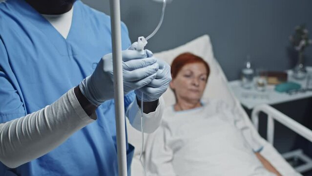 Tilting up of young Black male nurse wearing scrub suit, gloves and eyeglasses adjusting dropper of mature red-haired Caucasian woman who lying in hospital bed