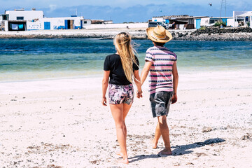 Fototapeta na wymiar Couple of young tourist walking together barefoot at the sandy beach. Concept of summer holiday vacation people. Boy and girl holding hands and blue ocean in background. Travel lifestyle
