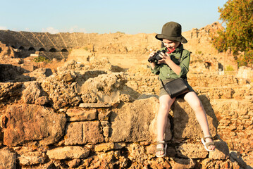 Boy archeologist in khaki clothes sitting on ruins and looking into the binoculars against ancient amphitheater on background. Diverse, non-traditional job concept - obrazy, fototapety, plakaty