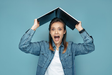 Excited young woman in jeans shirt holding computer or laptop over head in front of blue background.